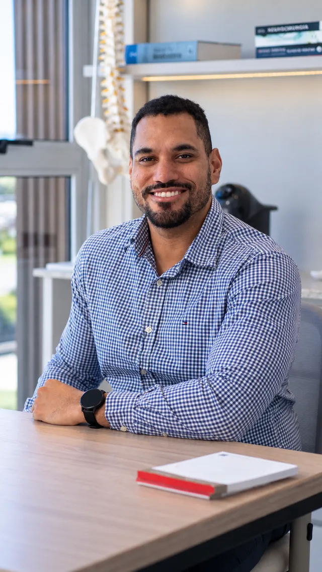 Dr. Elson Shockness sorrindo, de camisa xadrez, ao lado da janela do consultório